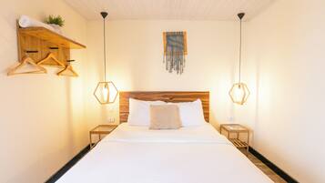 Una habitación de hotel con cabecera de madera, cama con ropa blanca, dos mesitas de noche con lámparas y un estante de madera con una toalla y una planta.