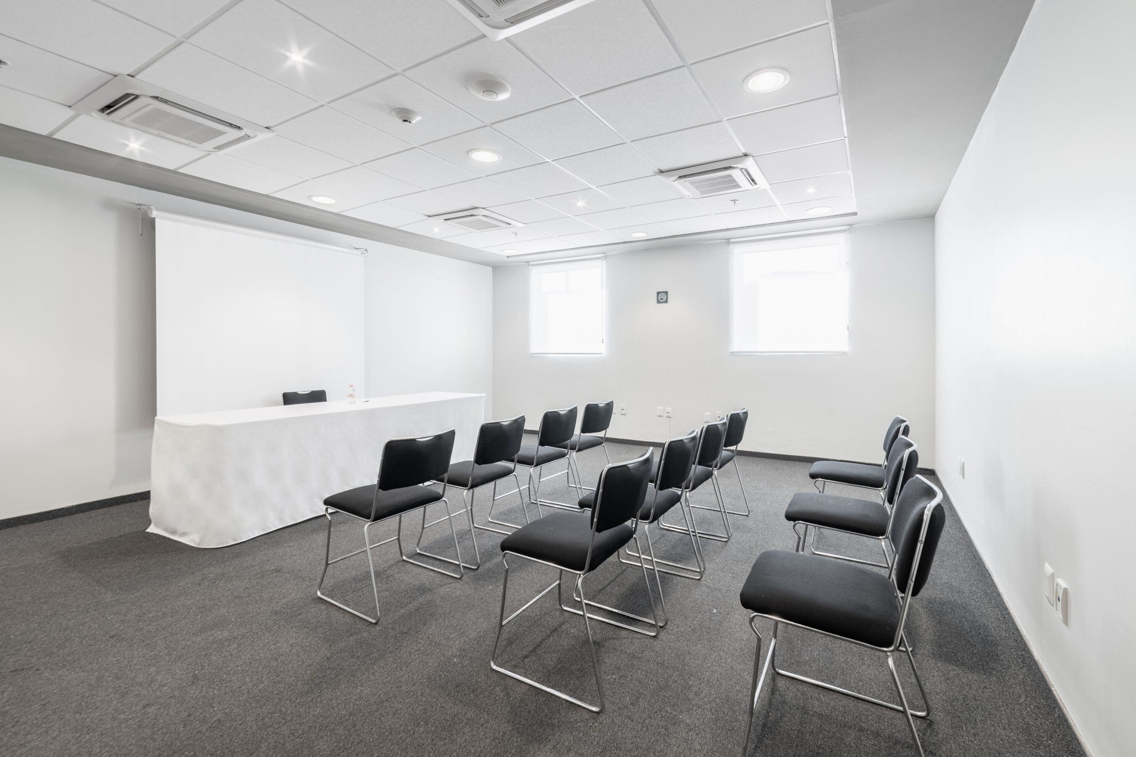 Un salón de conferencias con una mesa blanca, sillas negras, una pantalla de proyector y ventanas amplias.