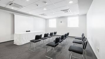 Un salón de conferencias con una mesa blanca, sillas negras, una pantalla de proyector y ventanas amplias.