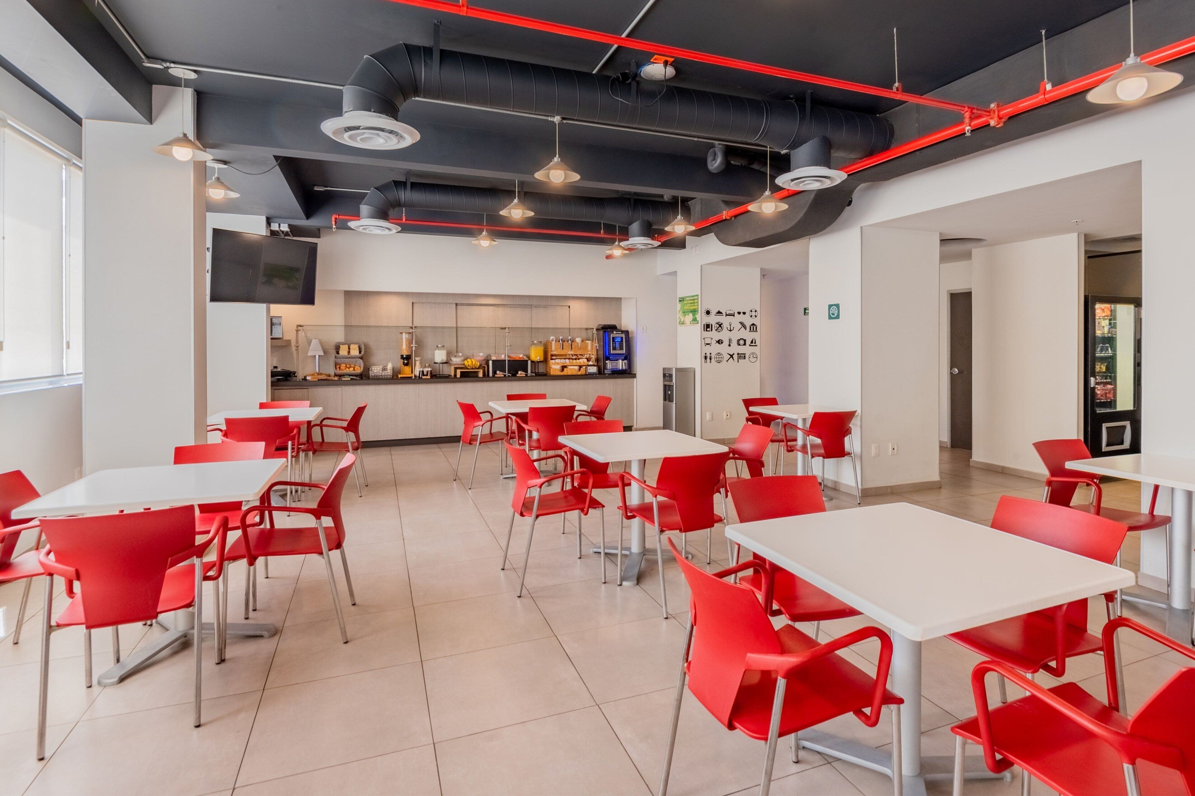 A modern dining area with red chairs and white tables, a bar area in the background, and a clean, industrial ceiling design.