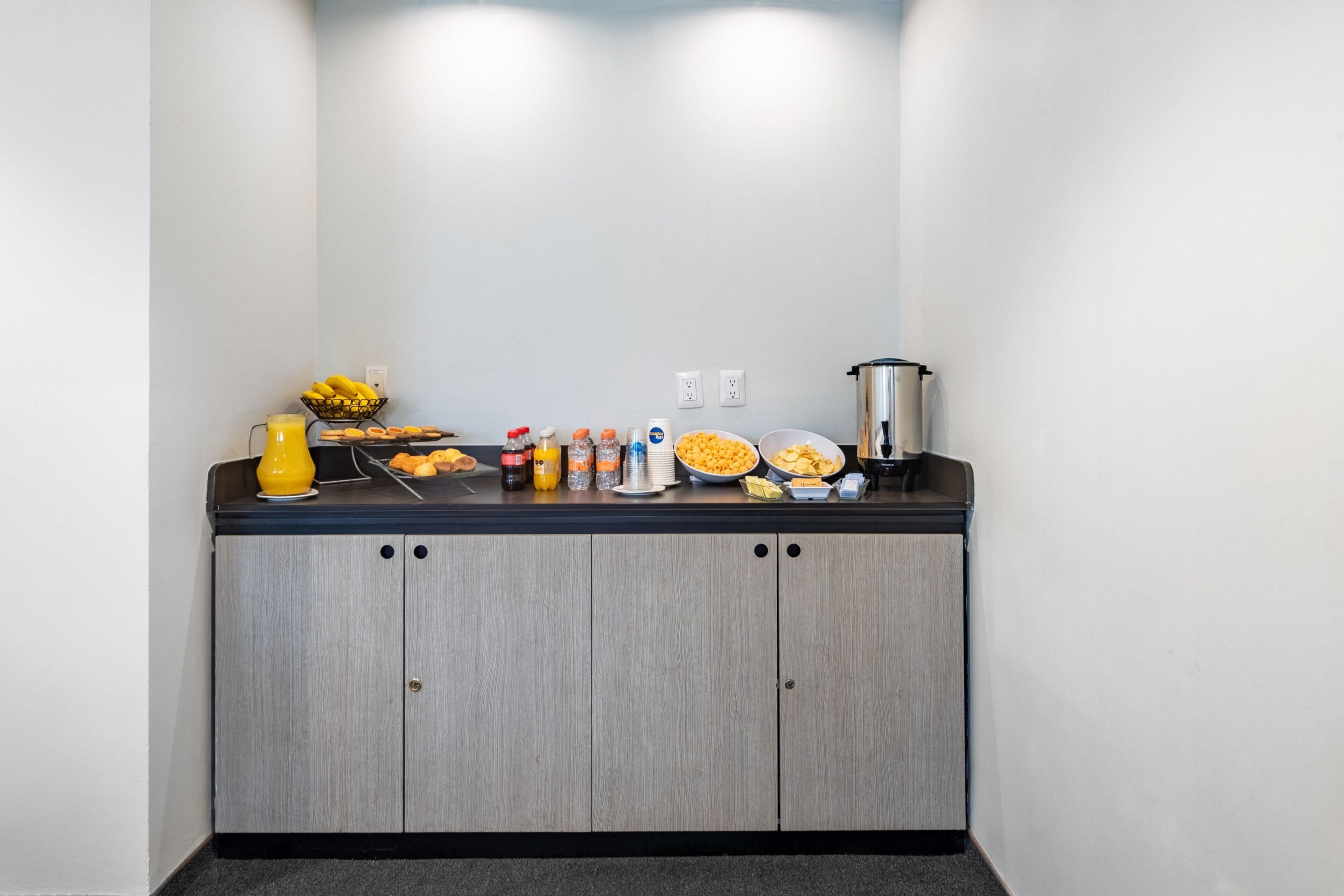 Una kitchenette con encimera negra, un florero amarillo, un cesto de plátanos, un tazón de cereal y una cafetera.