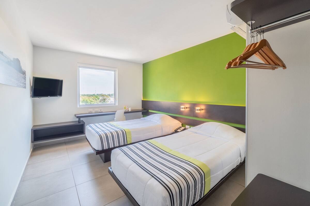Habitación de hotel con dos camas, un televisor, una ventana y una pared de acento verde.