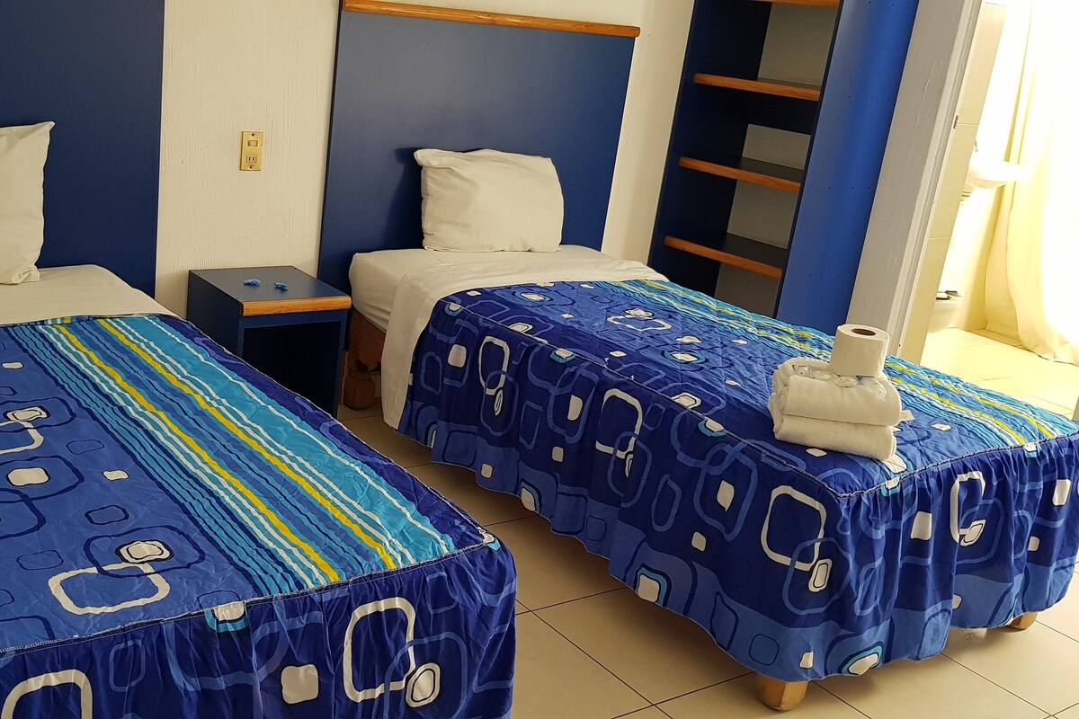 Une chambre d’hôtel avec deux lits, chacun recouvert d’une couvre-lit à motifs bleus et agrémenté d’oreillers blancs. Une petite table de chevet avec une lampe s’y trouve.