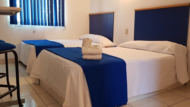 A hotel room with two beds, each with a blue bedspread and white pillows. There is a chair and a small table in the room.