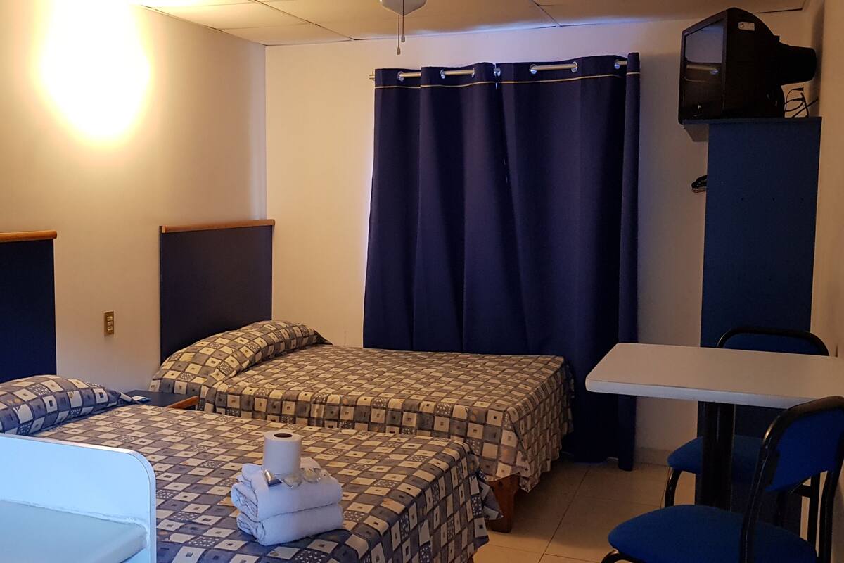 Une petite pièce, de taille modeste, comprenant deux lits, un bureau, une chaise et un téléviseur posé sur un meuble.