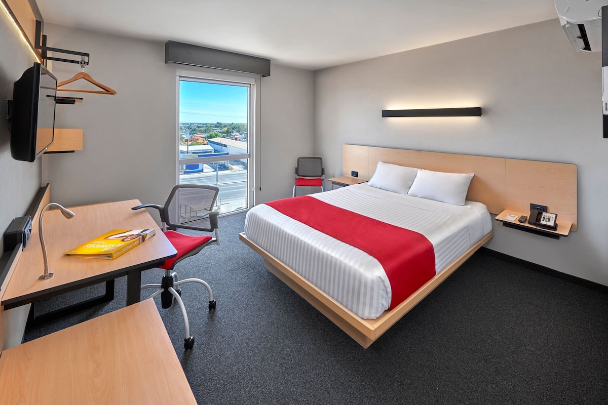 Habitación de hotel con cama, escritorio, silla, televisor montado en la pared y ventana con vista a la ciudad.