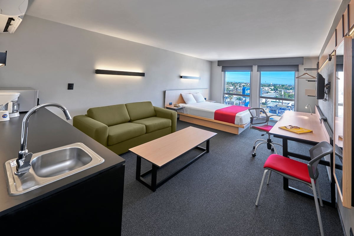 Una habitación de hotel moderna con kitchenette, un sofá verde, una cama con una colcha roja, un escritorio con silla y un ventanal con vistas a la ciudad.