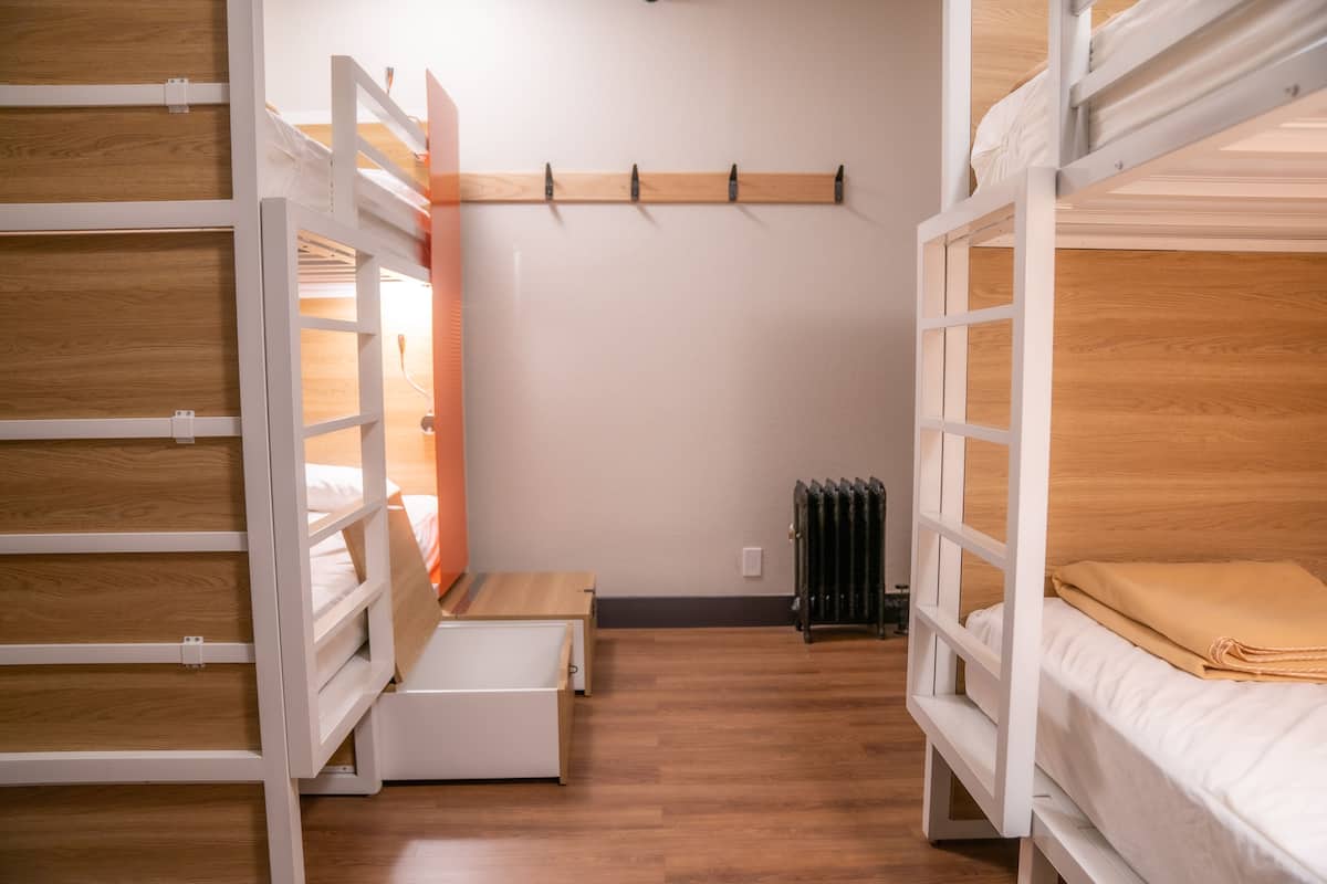 A dormitory room with bunk beds, a wooden floor, a radiator, and a wall-mounted coat rack.