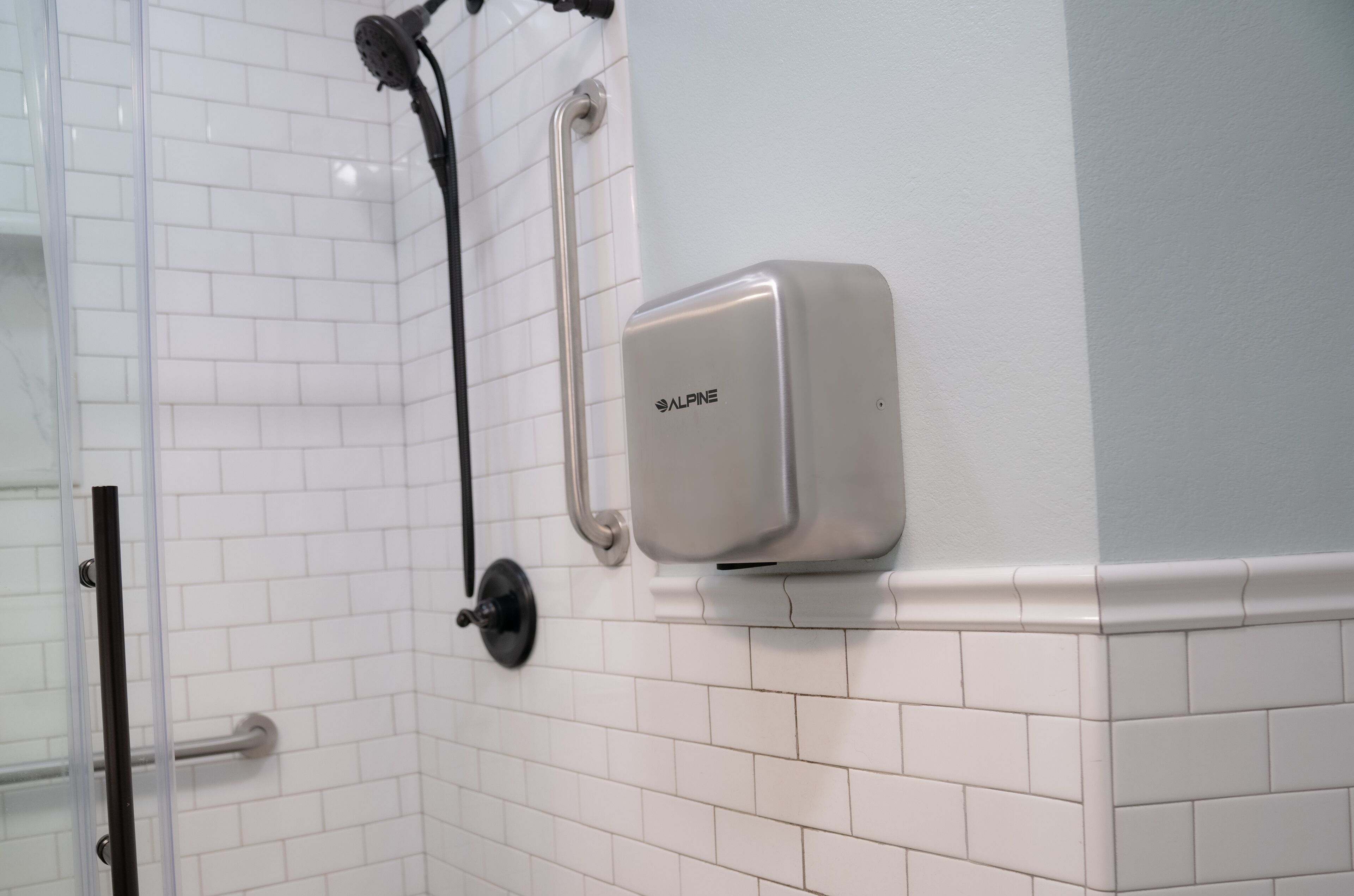 A bathroom with white tiled walls, a shower with a handheld showerhead, and a soap dispenser mounted on the wall.