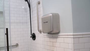 A bathroom with white tiled walls, a shower with a handheld showerhead, and a soap dispenser mounted on the wall.