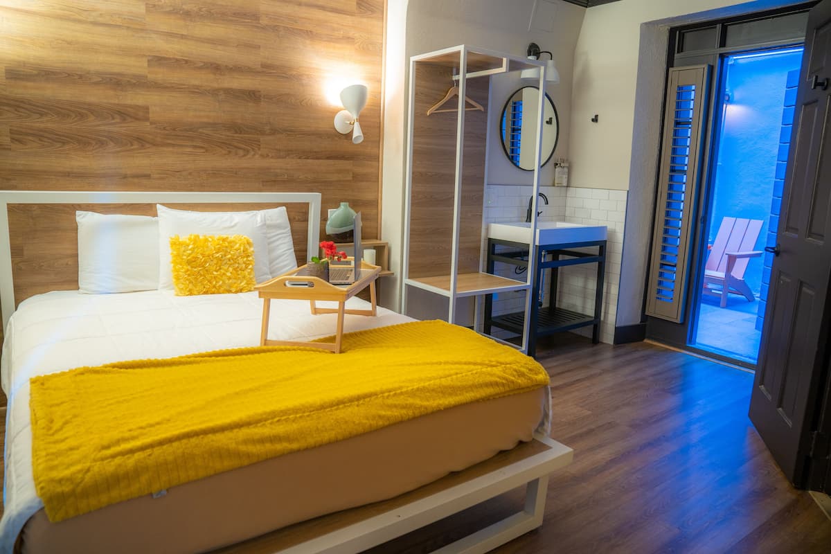 A hotel room with a bed, a bedside table, a mirror, a hanging coat rack, a bathroom with a sink and toilet, and a view of the outside.