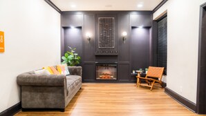 A living room with a grey sofa, a wooden floor, a fireplace, and a small table with a chair. - Amsterdam Hostel (San Francisco)