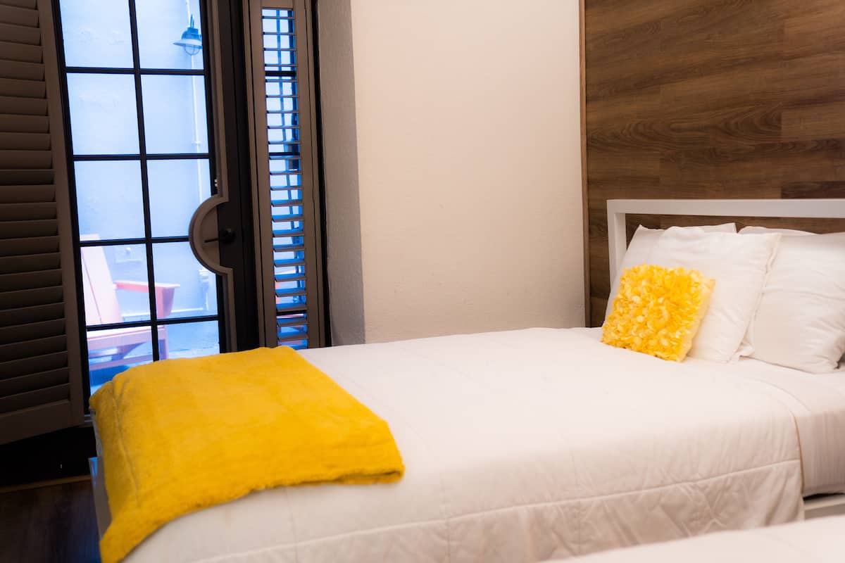 A hotel room with a bed, a yellow throw blanket, and a view through a window with blinds.