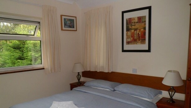 Ein ordentlich bezogenes Bett mit blauer Bettdecke, zwei Kissen und zwei weißen Handtüchern. Ein Fenster mit Vorhängen, ein gerahmtes Bild an der Wand und eine Lampe auf dem Nachttisch sind zu sehen.