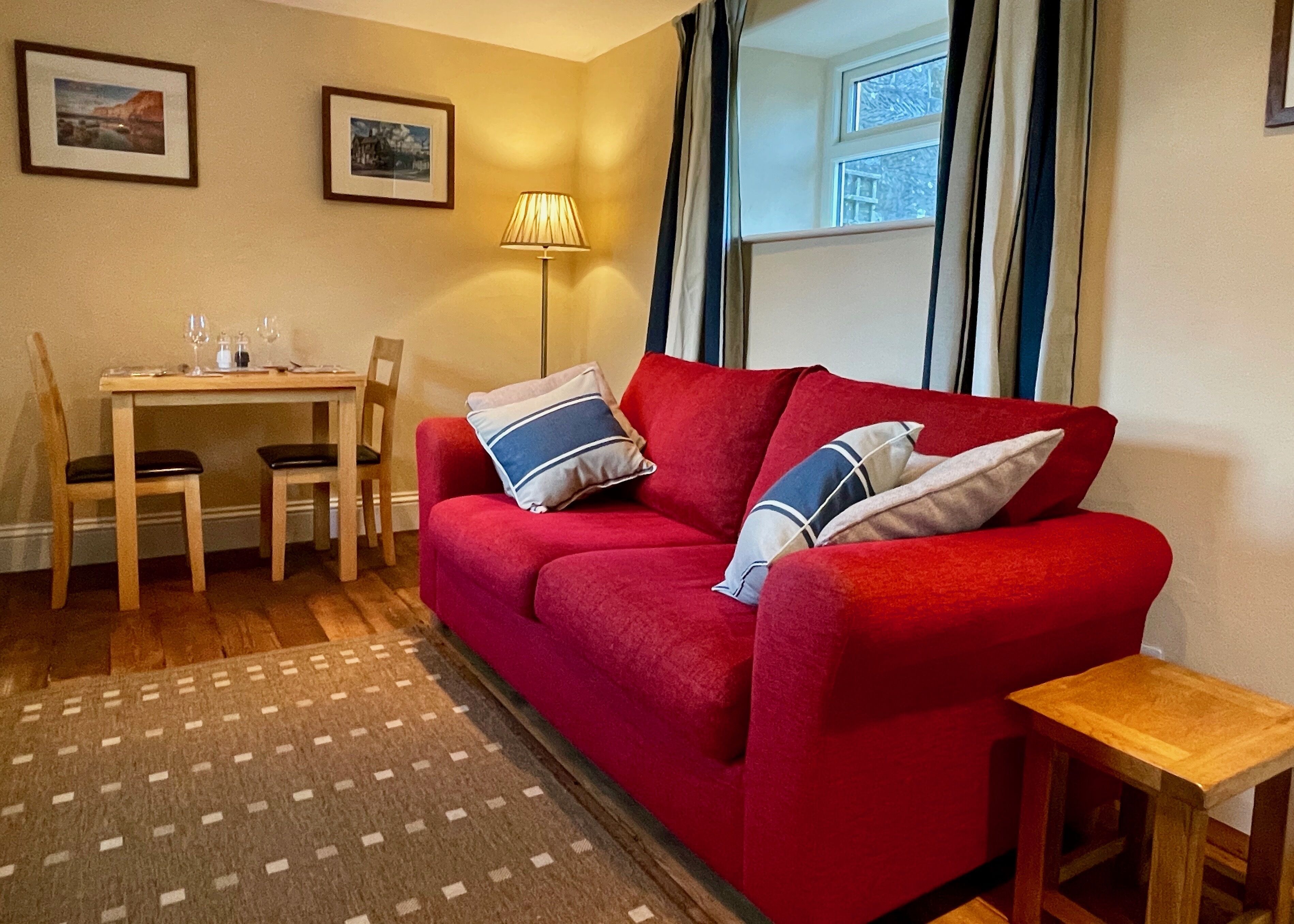 A red sofa with two pillows, a wooden table with chairs, a lamp, and two framed pictures on the wall.