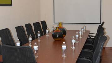 Un salón de conferencias con una larga mesa, sillas negras, botellas de agua y una pantalla para proyector.