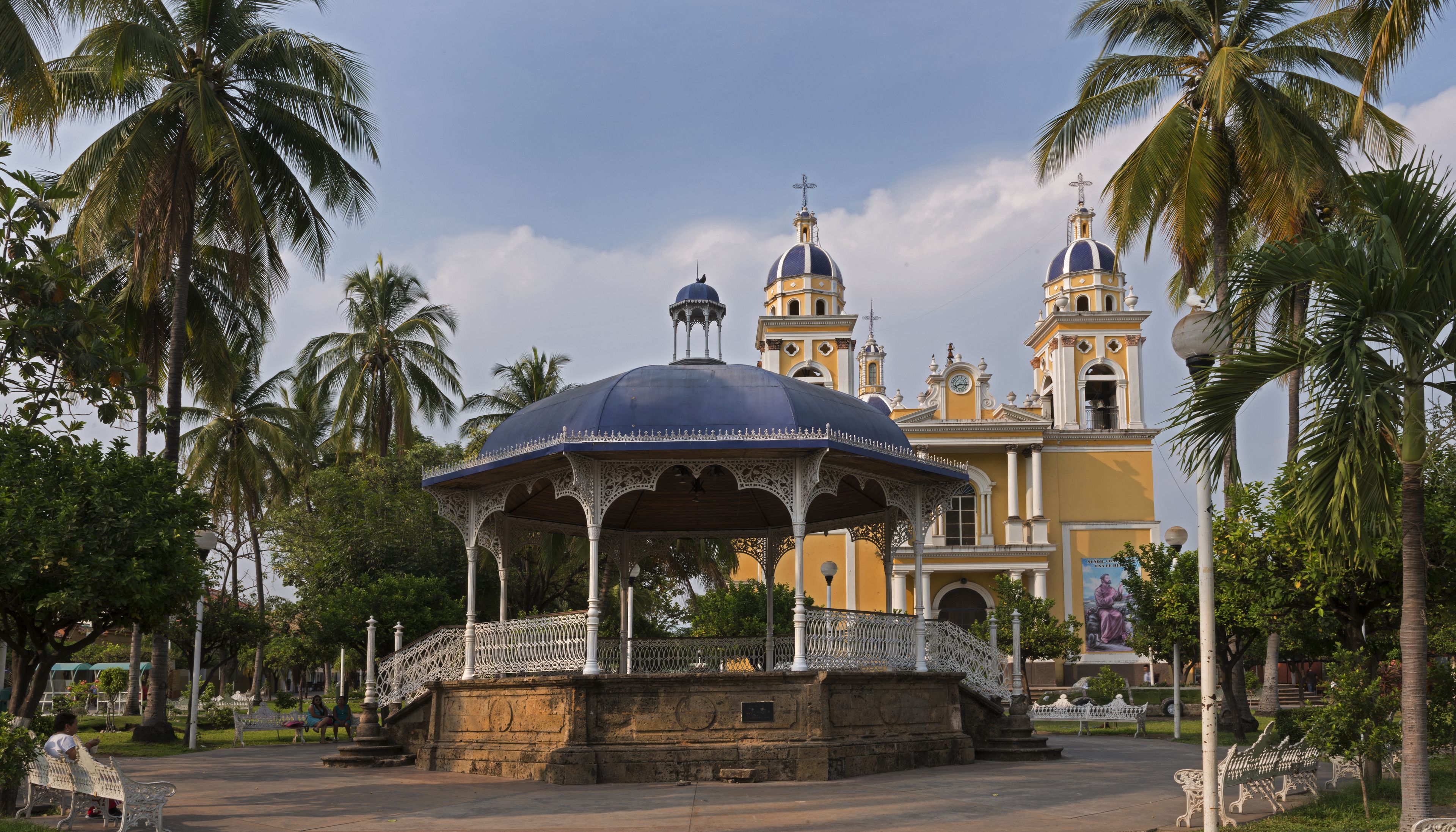 Un quiosco con barandillas blancas ornamentadas, base de piedra y cúpula azul, rodeado de palmeras y un edificio amarillo con cúpulas y pináculos al fondo.