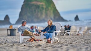 Dos personas sentadas en sillas blancas sobre una playa arenosa, con una gran formación rocosa al fondo.