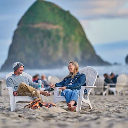 Two people sitting on white chairs on a sandy beach with a large rock formation in the background.
