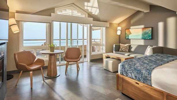 Un hotel moderno con una cama grande, un área de descanso con mesa y sillas, vista al mar a través de amplios ventanales y un cuadro en la pared.