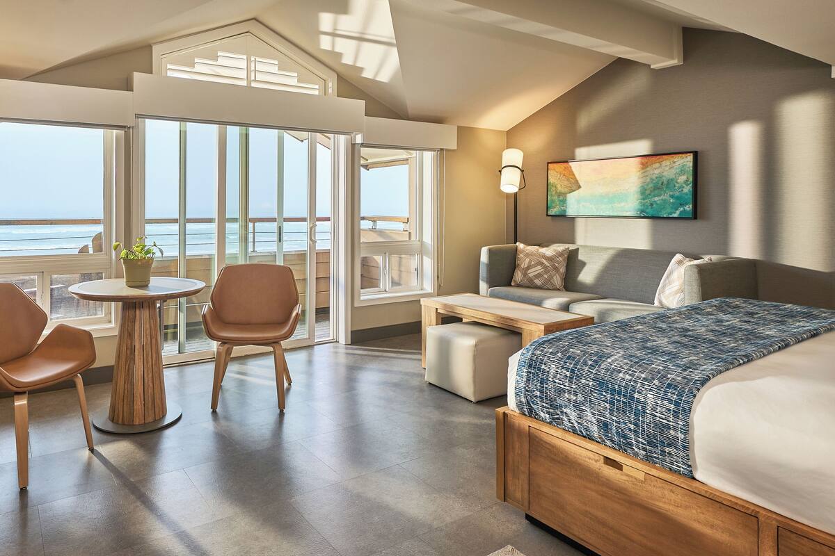 A modern hotel room with a large bed, a seating area with a table and chairs, a view of the sea through large windows, and a painting on the wall.