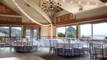 Un salón de banquetes con mesas redondas dispuestas para comer, amplias ventanas con vistas al océano y una lámpara de cristal.