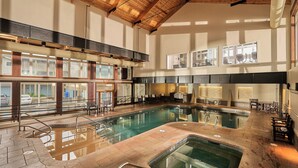 Una amplia piscina interior con techo de madera, varios niveles con barandillas de cristal y zonas de estar alrededor de la piscina.