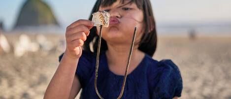 A person holding a marshmallow on a stick, with a beach and a structure in the background.