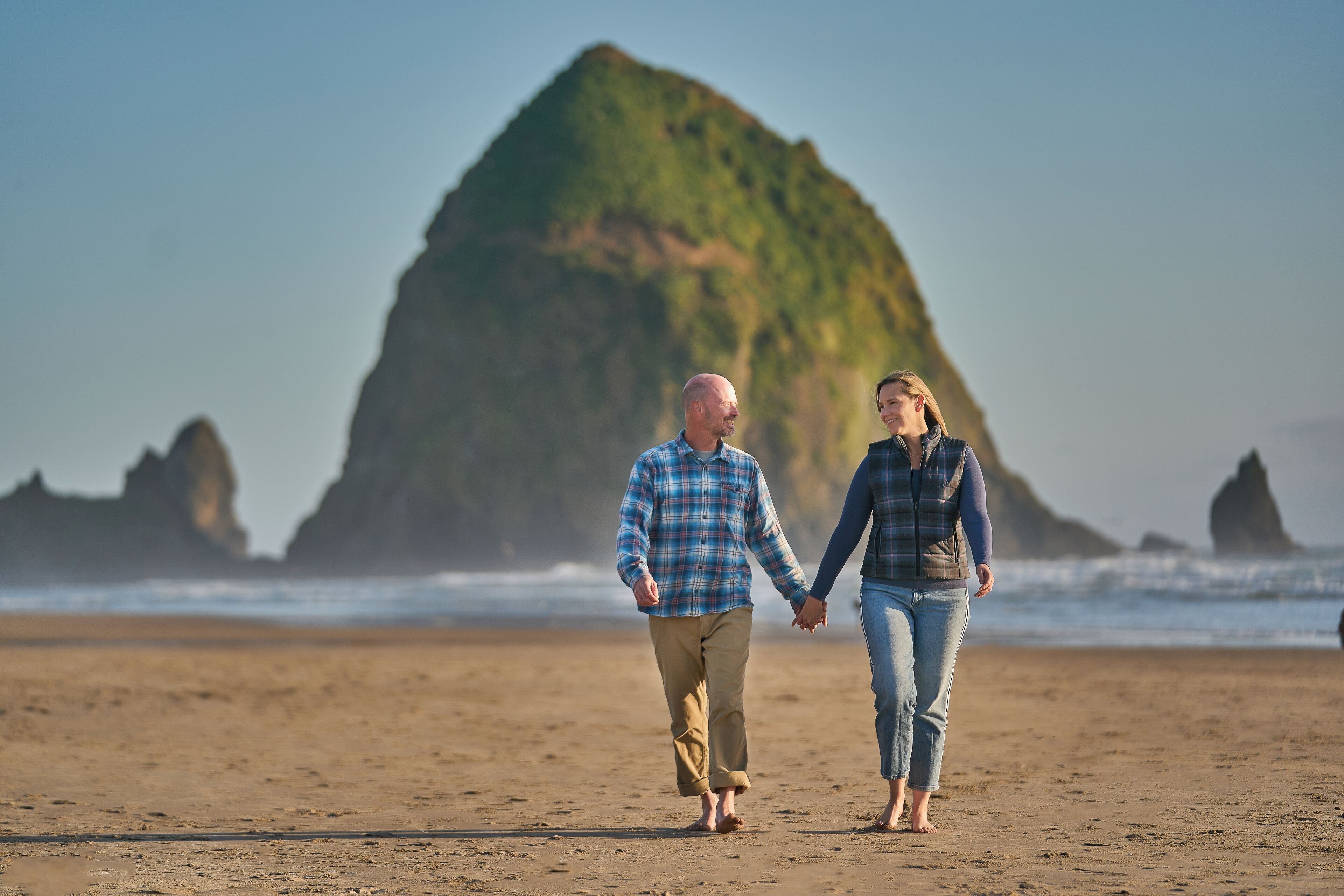 Two people walking on a sandy beach with a large rock formation in the background.