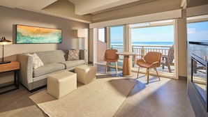 Una habitación de hotel moderna con sofá, pufs, una mesita y sillas. Hay un ventanal con vistas al océano.