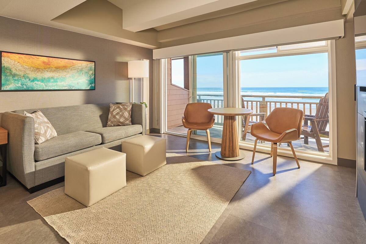A modern hotel room with a sofa, ottomans, a small table, and chairs. There is a large window with a view of the ocean.