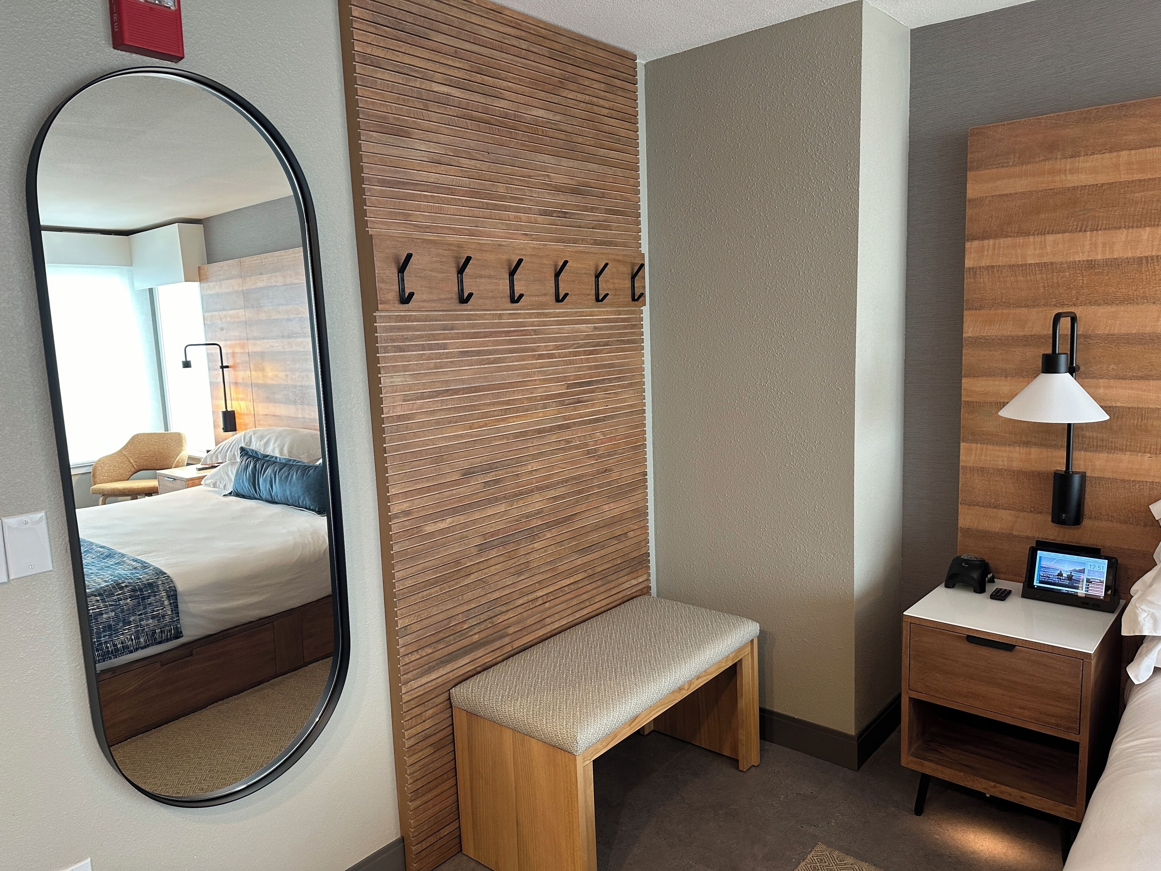 A hotel room with a large mirror, a wooden panel wall, a bench, a bedside table with a lamp, and a digital clock.