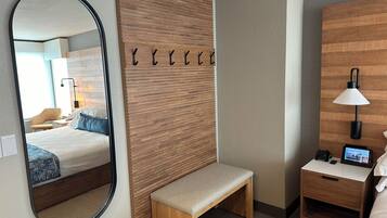 A hotel room with a large mirror, a wooden panel wall, a bench, a bedside table with a lamp, and a digital clock.