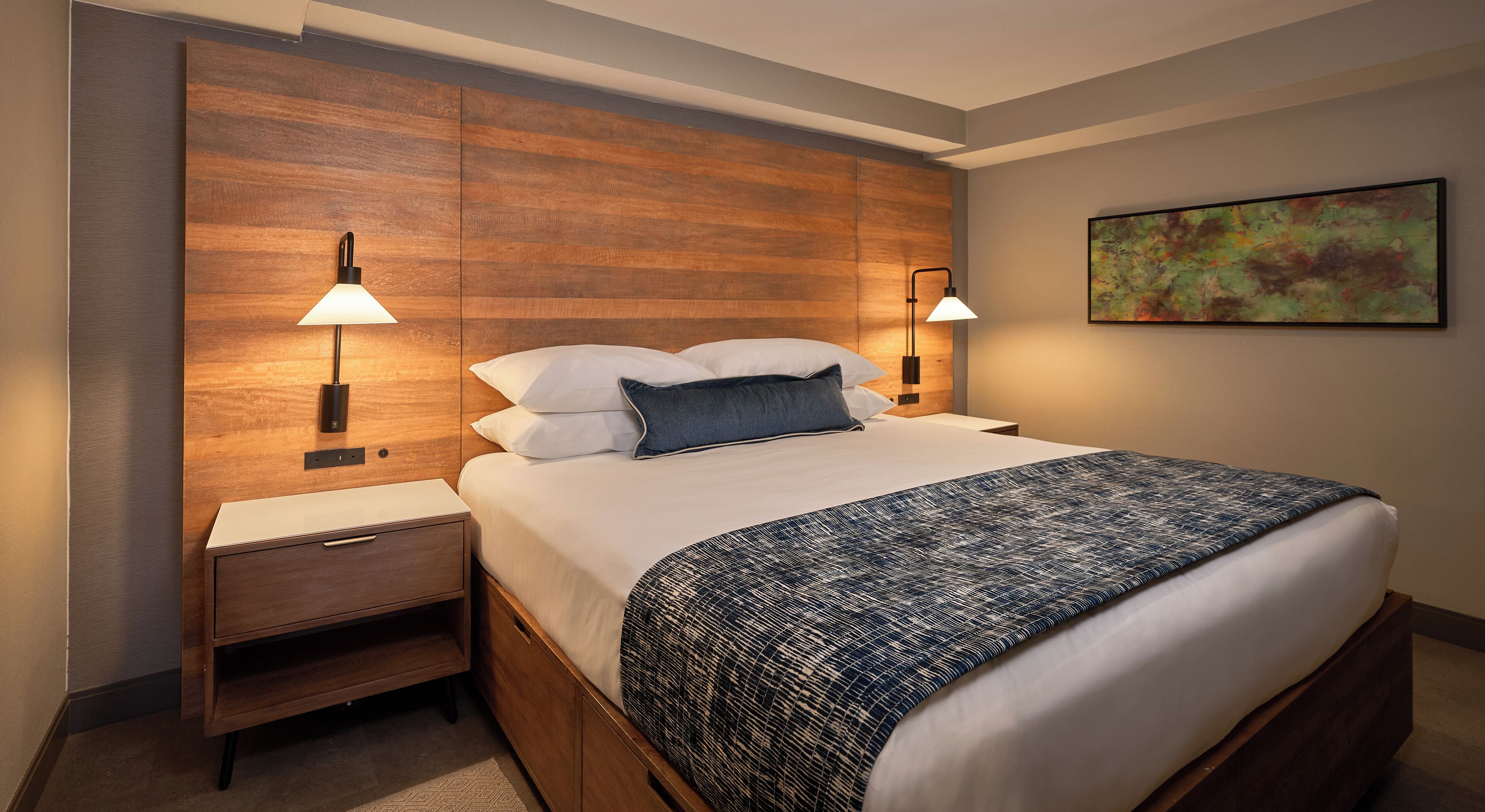 Ein Hotelzimmer mit einem hölzernen Kopfteil, zwei Nachttischlampen, einem Bett mit weißen Bettwäsche und einer gemusterten Tagesdecke, einem Nachttisch und einem Bild an der Wand.