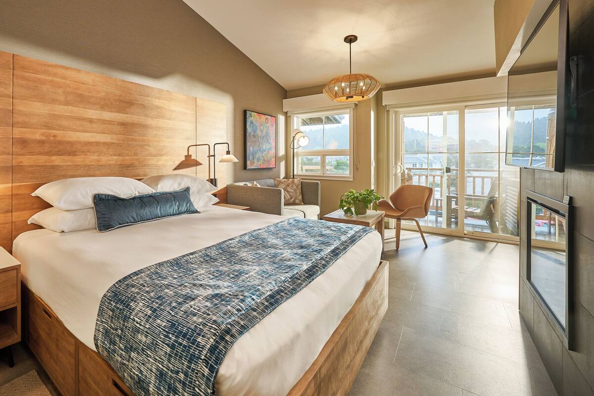 A modern hotel room with a wooden headboard, a large bed with a blue patterned throw, a nightstand, a desk with a chair, and a balcony with a view.