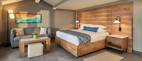 Un dormitorio moderno con cabecera de madera, cama con ropa blanca, sofá gris, mesita de noche de madera y lámpara de pie.
