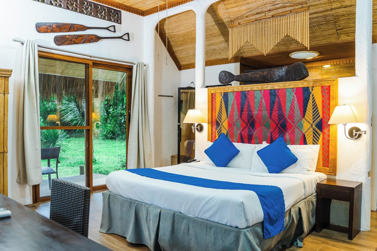 Une chambre spacieuse avec un grand lit, une vue sur le jardin grâce à de grandes portes vitrées et un plafond en bois.