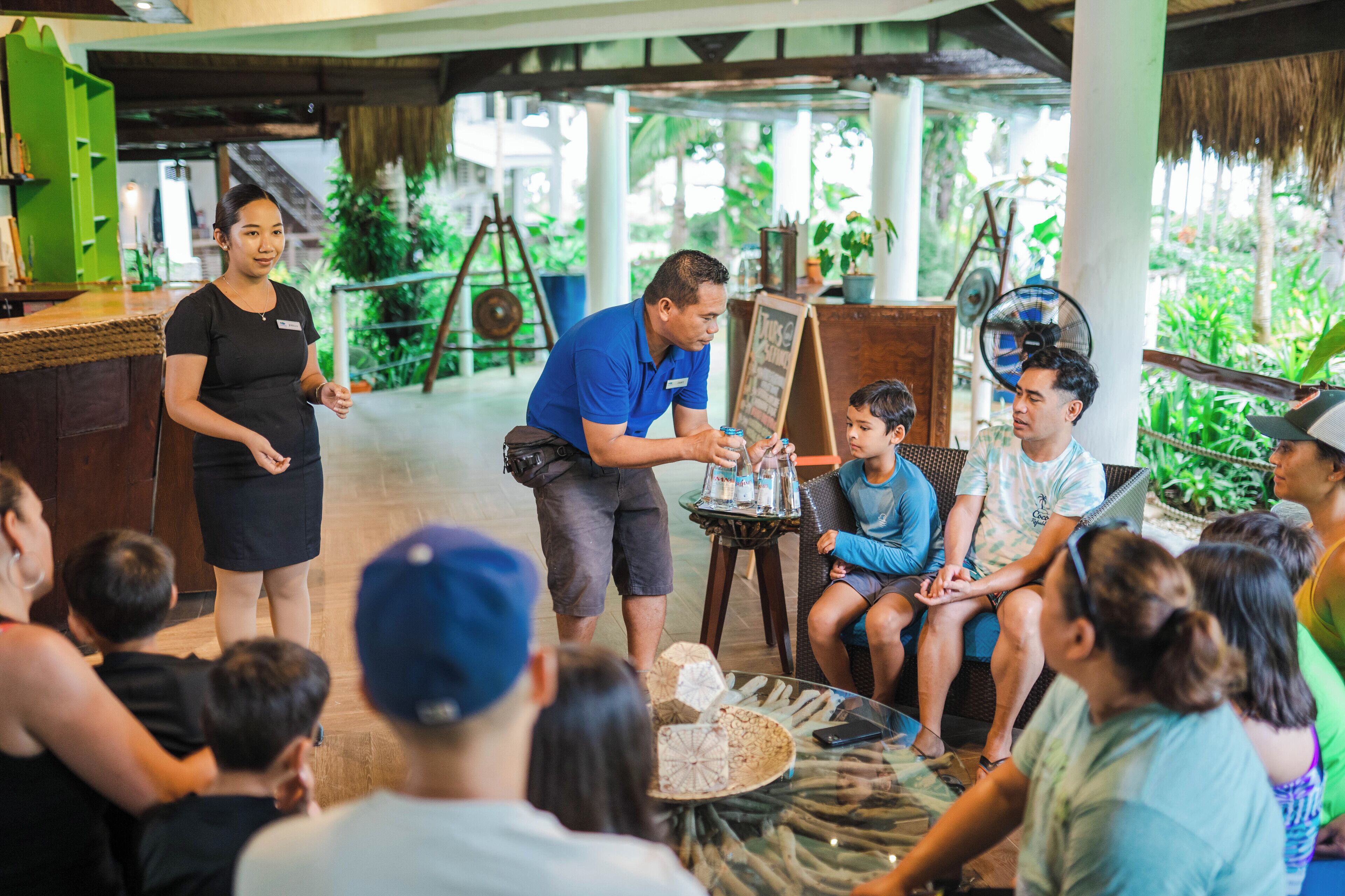 A group of people seated around a table with a man presenting in a casual setting with a thatched roof and greenery in the background.