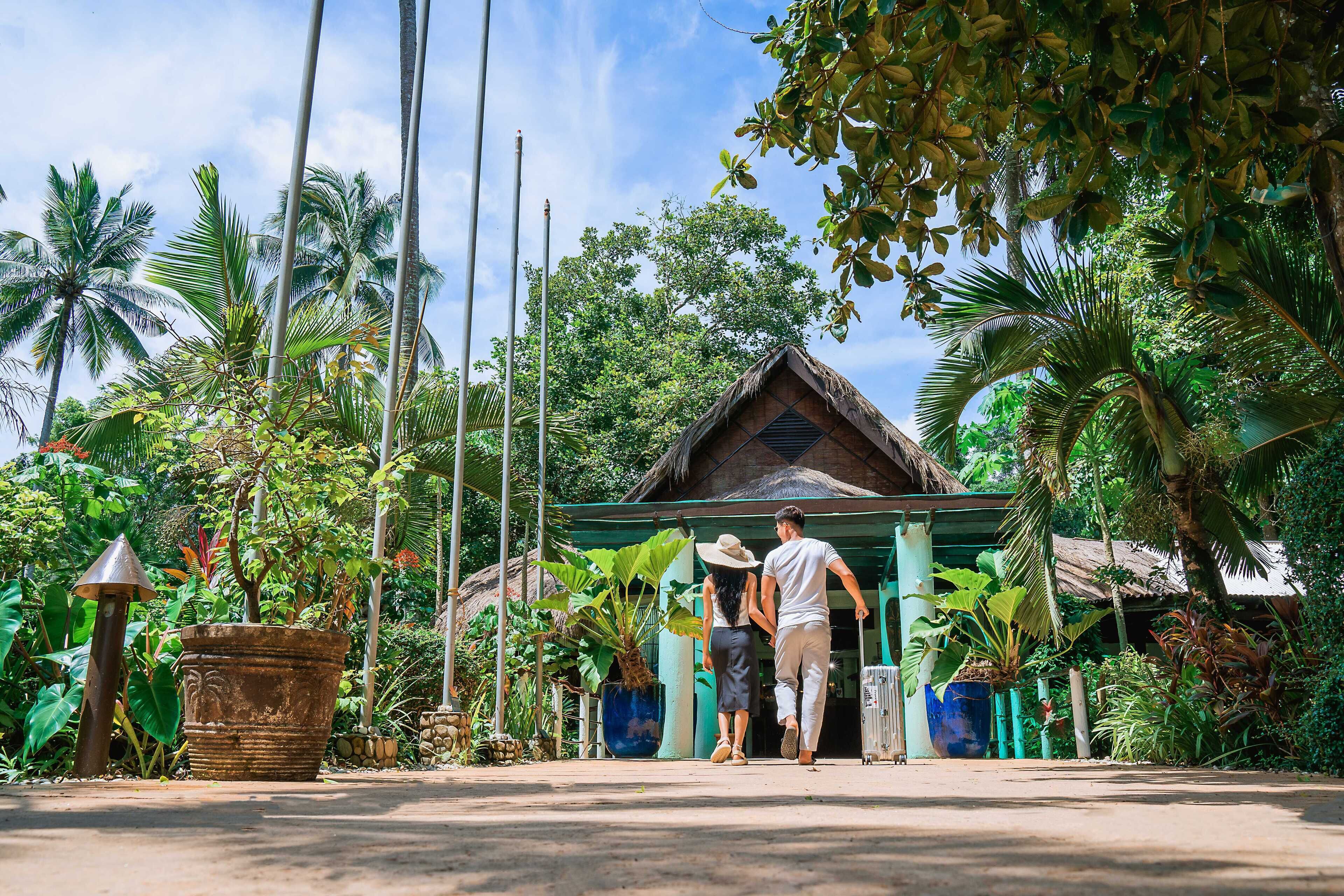 A couple walking towards a traditional thatched-roof building surrounded by lush tropical vegetation.