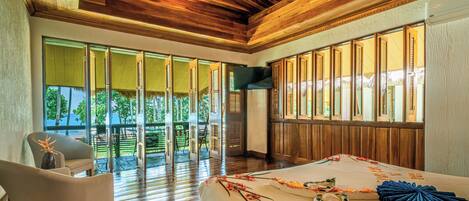 A spacious room with a wooden ceiling, large sliding glass doors leading to an outdoor area, a bed with floral decorations, and a chair.