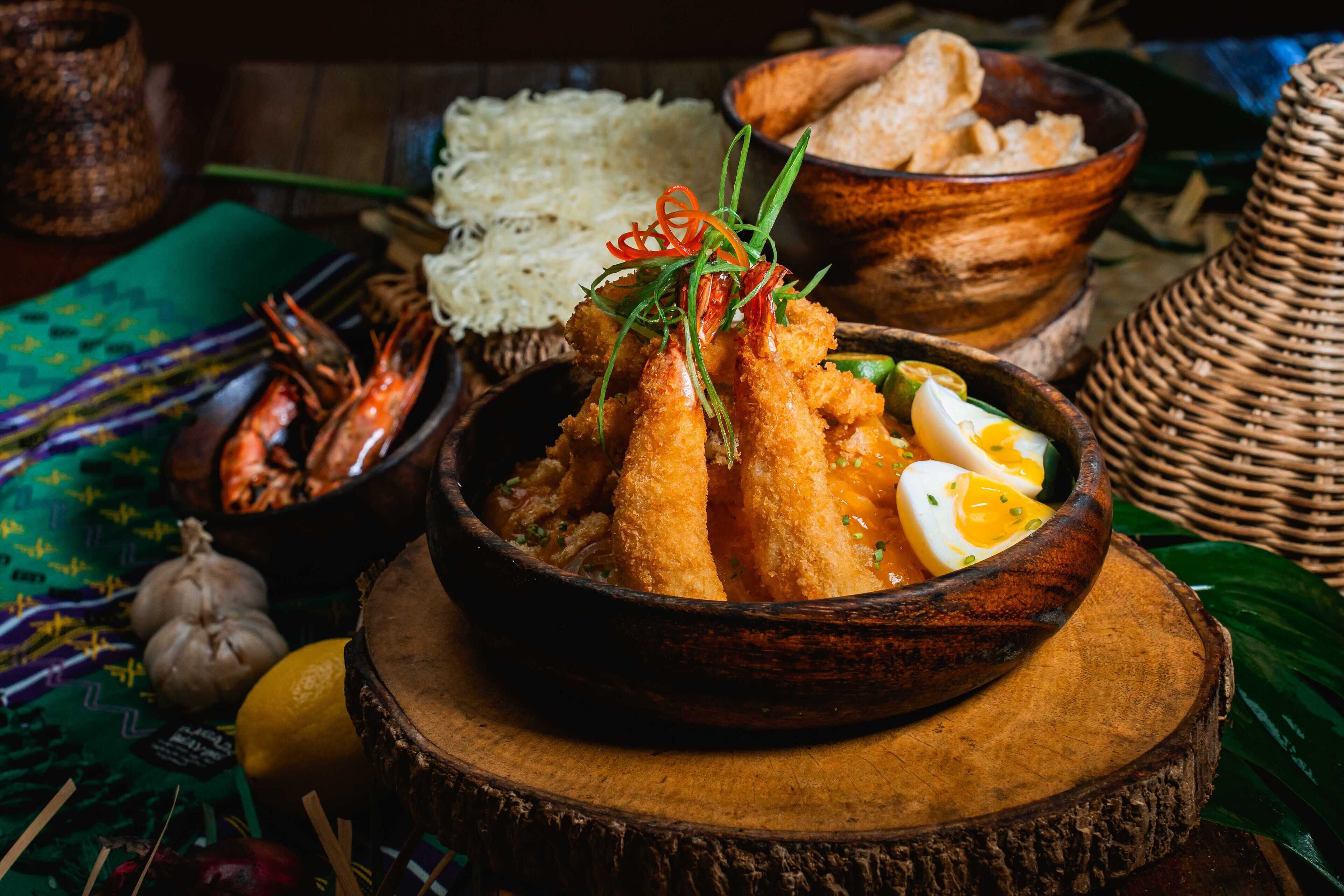 A wooden bowl with fried food garnished with herbs, surrounded by bowls of rice and other dishes.