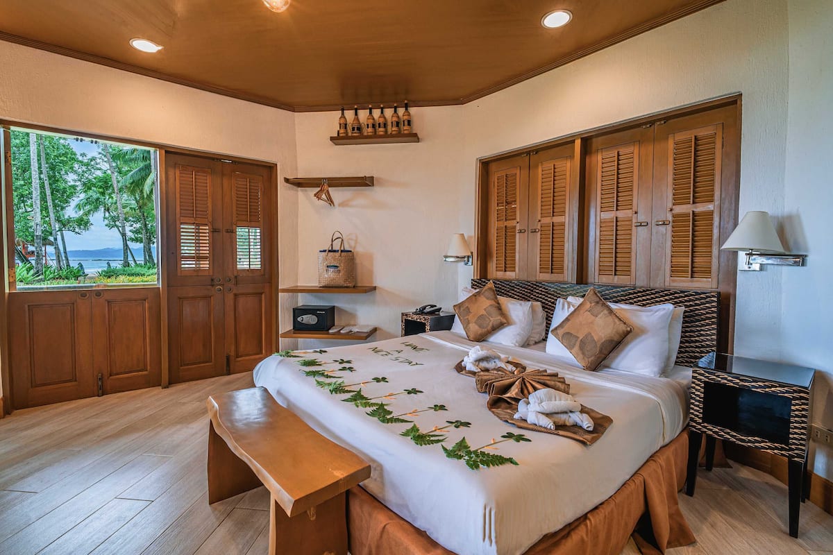 Une chambre d’hôtel avec un grand lit, des volets en bois, une vue sur des palmiers et un banc en bois.