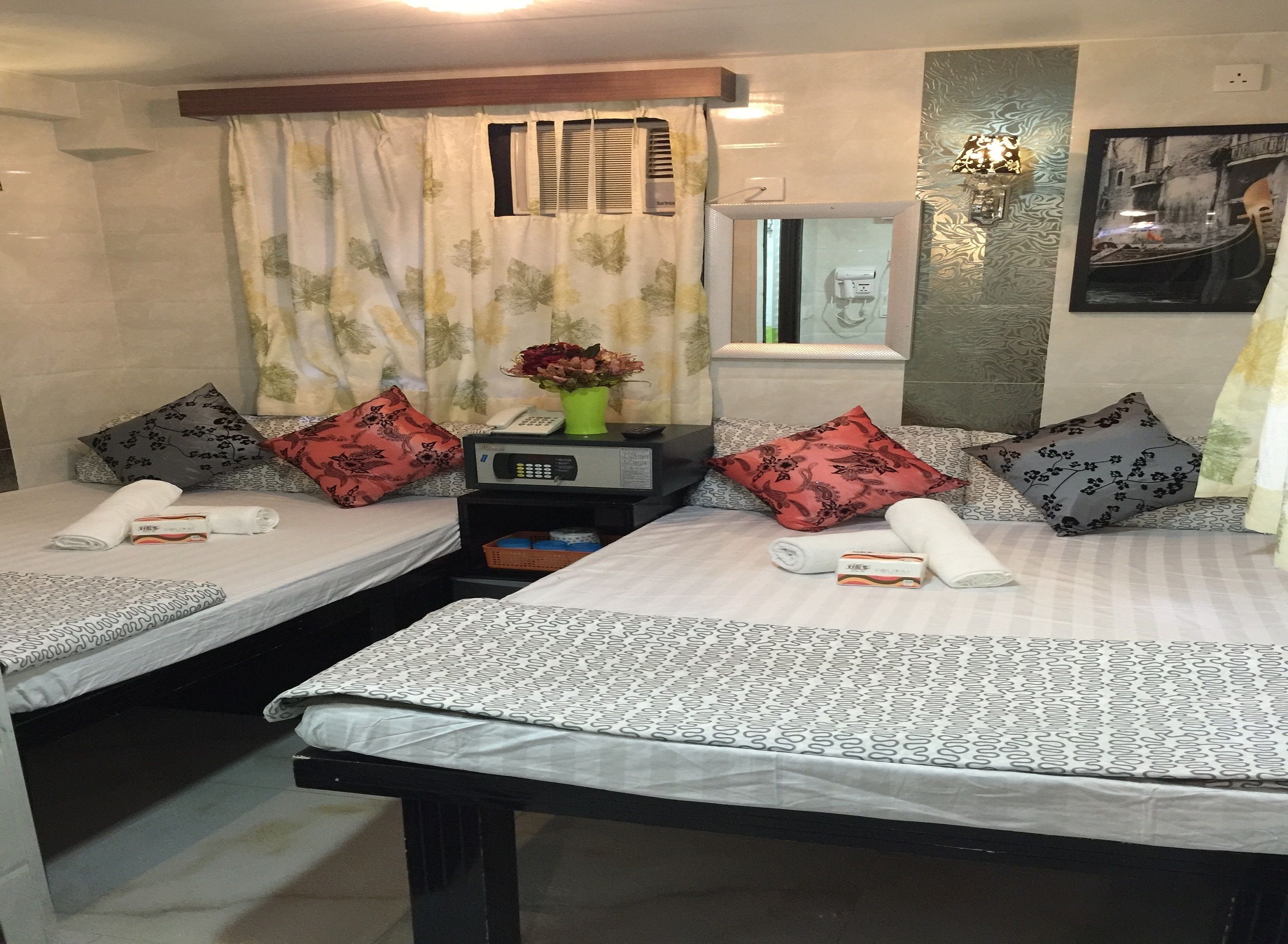 Une chambre d’hôtel avec deux lits, chacun recouvert d’une couvre-lit à motifs, agrémenté d’oreillers et de serviettes soigneusement pliées. On y trouve une table de chevet avec un téléphone et un petit vase avec des fleurs. Une fenêtre avec des stores et un luminaire fixé au mur sont visibles.