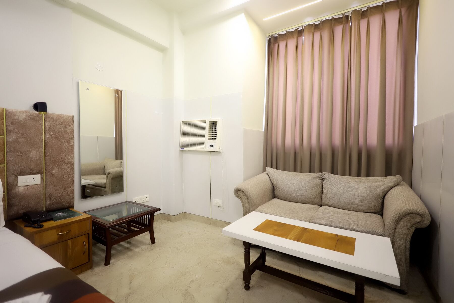A hotel room with a sofa, a small wooden table, a mirror, and a wall-mounted air conditioner.