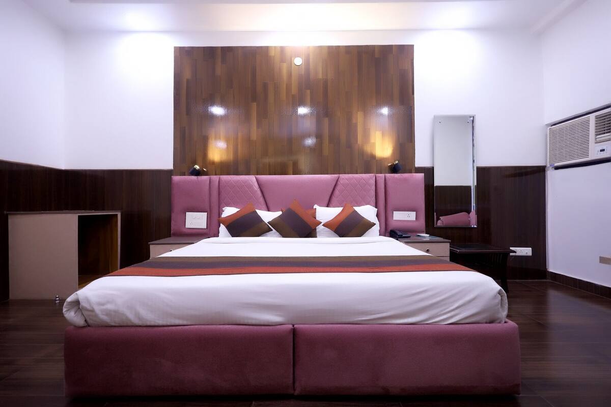 A modern hotel room with a large bed, a wooden headboard, a bedside table, a mirror, and a built-in air conditioning unit.