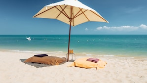 Une plage avec un parasol blanc, deux serviettes et une chaise sur le sable.