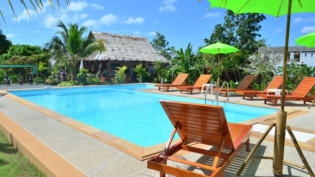 Una piscina azul cristalina con sillones de madera y sombrillas verdes, rodeada de vegetación tropical y un edificio con techo de paja.