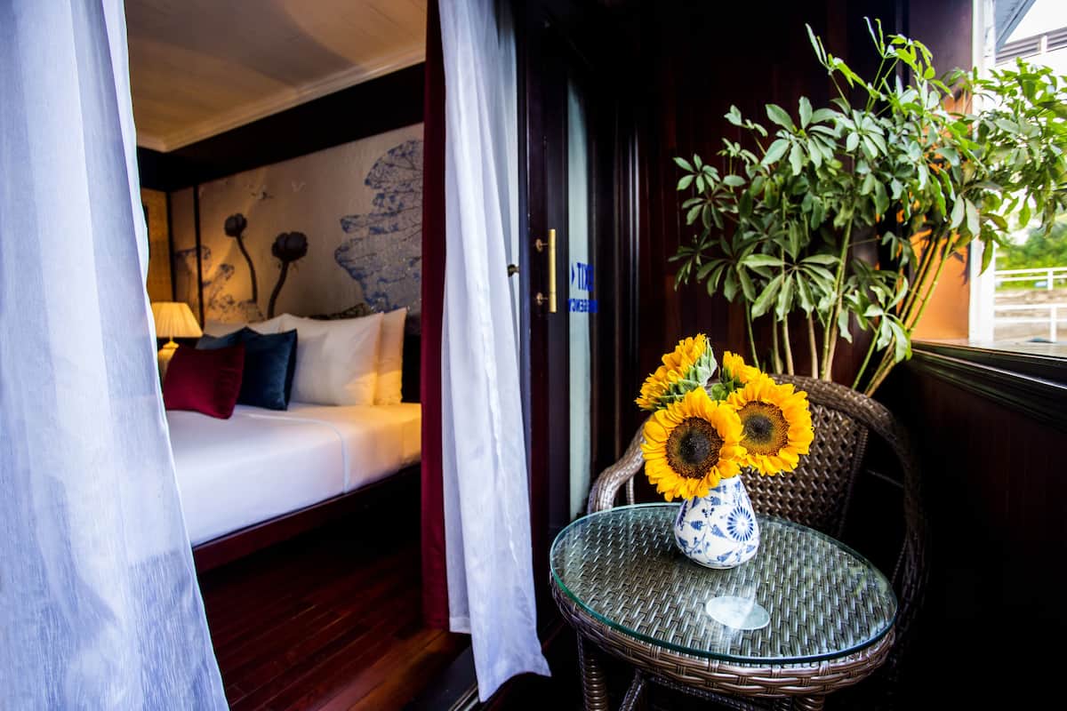 A room with a bed, a glass table with a vase of sunflowers, and a wicker chair.