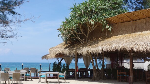 Un restaurante frente a la playa con techos de paja, muebles de mimbre y vista al océano.
