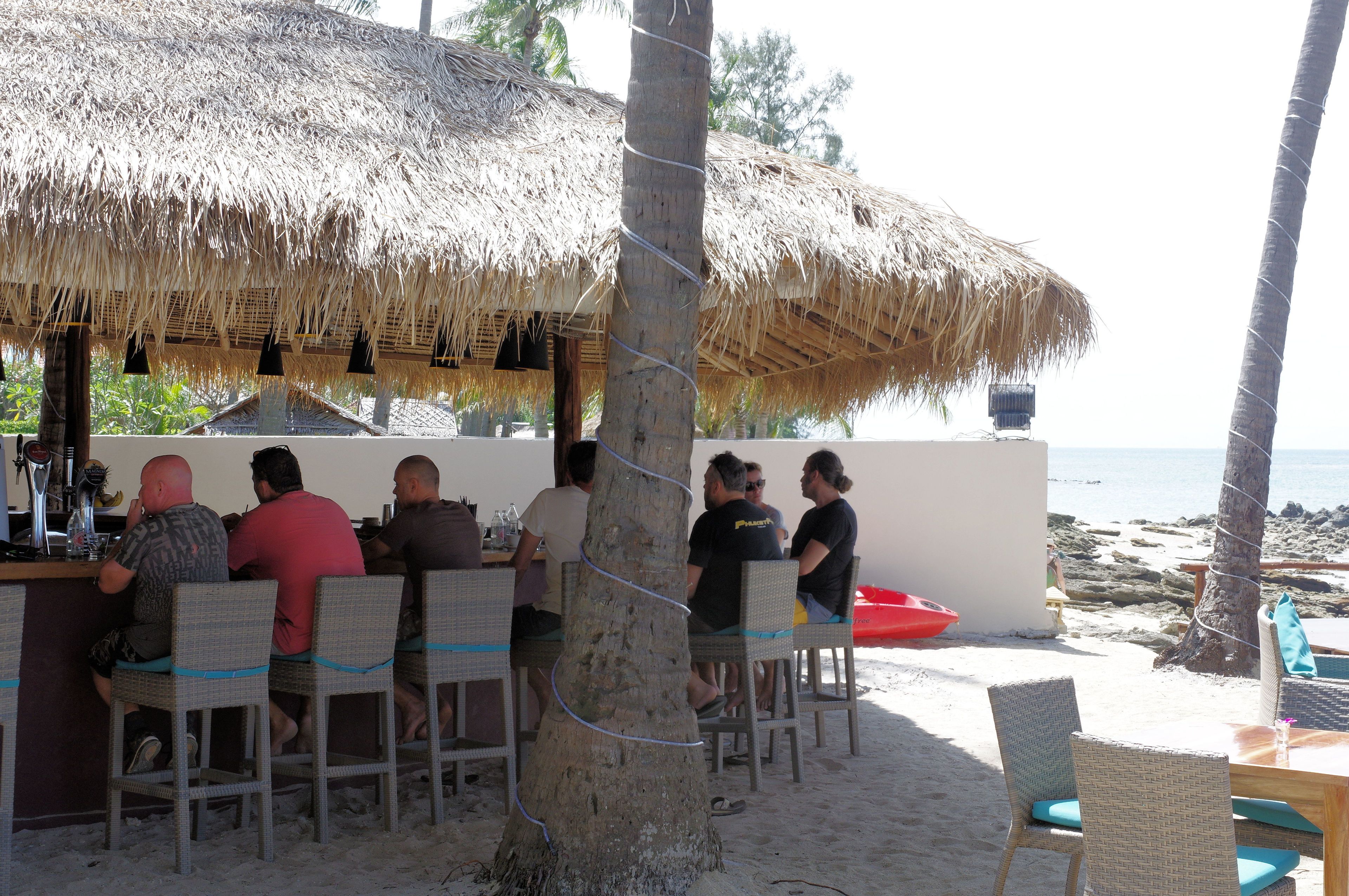 Un restaurante frente al mar con sombrillas de paja, sillas de mimbre y vistas al océano.